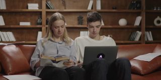 两个20多岁的学生在写文章，女性在书上听写信息，男性在笔记本电脑上打字。学生们在沙发上进行团队合作。