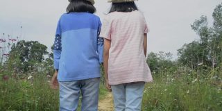 亚洲妇女和她的女儿戴着草帽，手牵手走在宇宙花田在夏天。后视图跟随镜头。