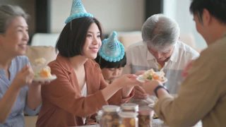 亚洲华人多代同堂庆祝生日，孙子戴着派对帽在客厅吃生日蛋糕视频素材模板下载