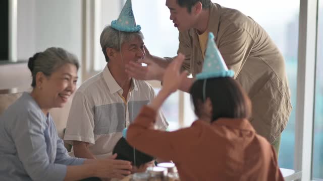 亚洲华人男子帮助他的父亲戴上派对帽，准备在家里的客厅庆祝生日