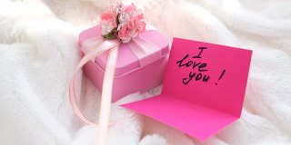粉红色的礼盒和纸条，上面写着“我爱你”，粉红色的明信片，上面盖着白色毛茸茸的毯子。温馨的卧室细节，给爱人的早晨