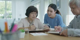 迷人的年轻老年亚洲公民夫妇坐在家里水彩画艺术品桌上与护士在心理健康治疗或亚洲老年人隔离活动生活方式感到快乐。