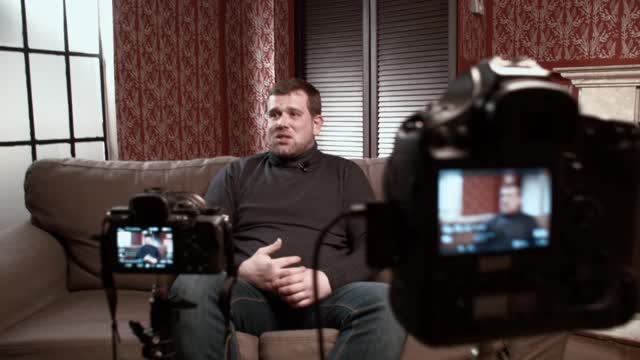 男博主接受采访时坐在沙发前摄像机前回答记者提问
