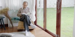 早上，退休的灰白头发的亚洲人爷爷坐在客厅看书，在家中自我隔离隔离。隔离或封锁期间在家活动的生活方式。
