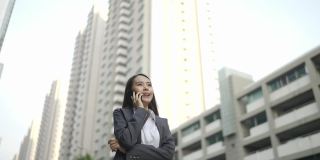 视频缓慢。亚洲女人，漂亮的女商人，白人，穿着黑色西装，在用智能手机聊天。在城市街道的一边