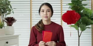 自拍视频通话中，一位迷人的年轻亚洲女士穿着红色的中国新年庆祝服，开心地微笑着交谈，看着拿着红包的手机。