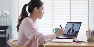 侧视图年轻的亚洲女性听观看数字课程在线学习员工技能在家庭办公室的笔记本电脑上提高技能。针对成人的商学院课程教育。