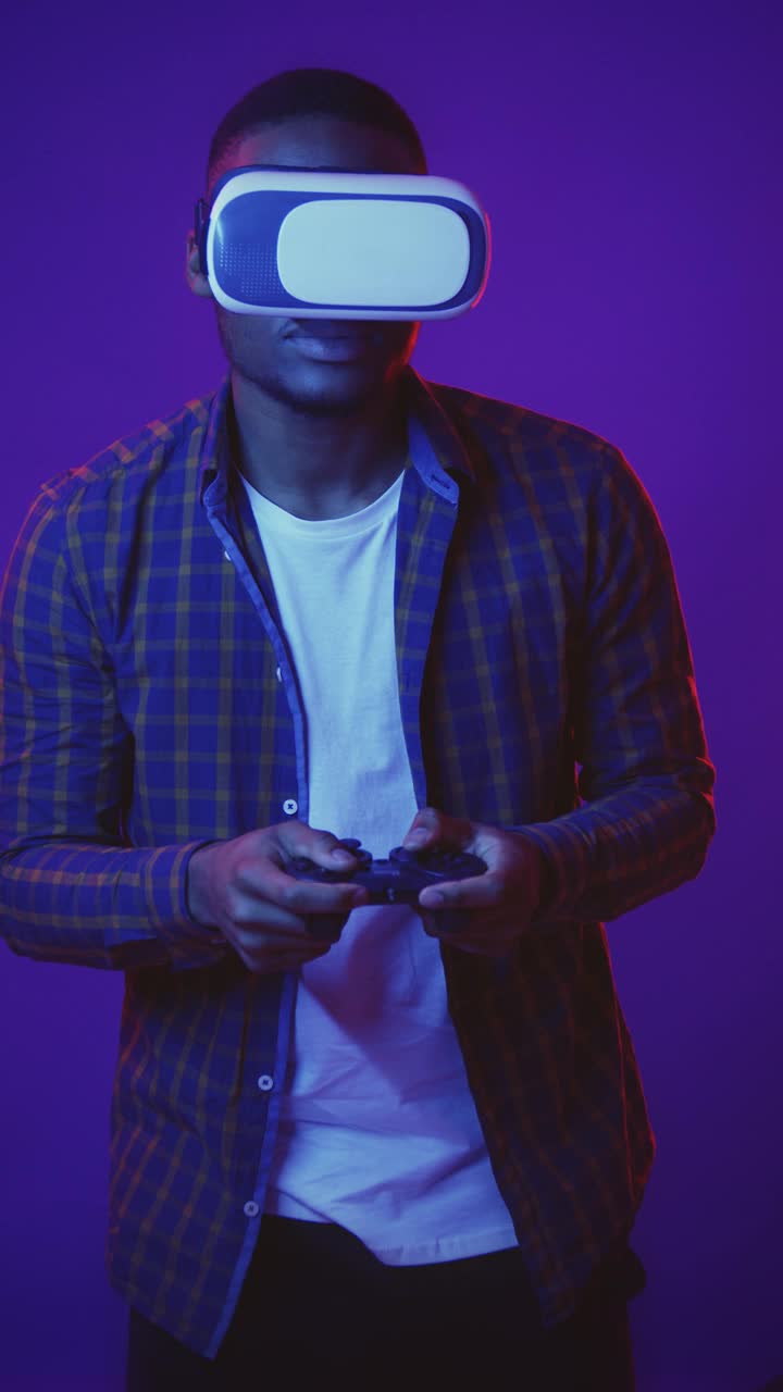 垂直工作室拍摄的年轻的非洲裔美国人戴着虚拟现实眼镜玩视频游戏与控制杆在霓虹灯