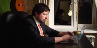 一个穿裤子、夹克、领带的男人晚上在家庭办公室的电脑前工作