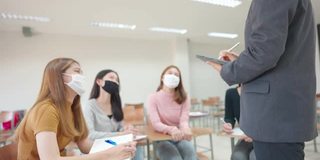 在大学教室里，一位老师用平板电脑给学生上课。学生戴口罩预防病毒新常态。