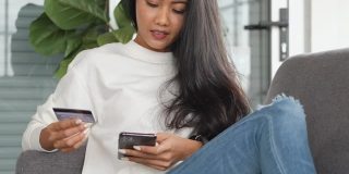 年轻的亚洲女性用智能手机和信用卡支付网上购物，坐在家里的沙发上。数字时代的生活方式