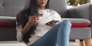 年轻的亚洲女性用智能手机和信用卡支付网上购物，坐在家里的沙发上。数字时代的生活方式