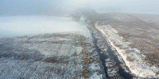 在一个霜冻和雾蒙蒙的早晨，一场雪之后，一条小河从荒地流向树木的无人机视图