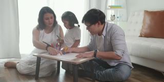 亚洲爸爸和妈妈一起在客厅教小女儿用彩色铅笔画和在纸书上画画