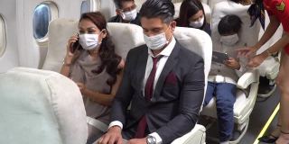 一对商务旅客戴着防护面具坐在飞机上一起聊天。在新冠肺炎大流行或新冠肺炎疫情期间乘坐飞机出行。女人用手机，男人用笔记本电脑