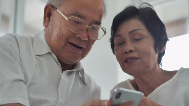 一对60-70岁的亚洲夫妇坐在养老院客厅的沙发上，一边聊天一边用智能手机看在线内容。高级技术的概念。
