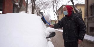 老人戴着防护口罩，用塑料刮冰器和扫把清理车窗上的积雪