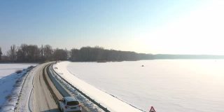 航拍的汽车行驶在冰雪覆盖的道路附近结冰的湖。冬季，白色越野车行驶在大坝路线上。飞过汽车，驶过河桥。风景优美的景观。俯视图