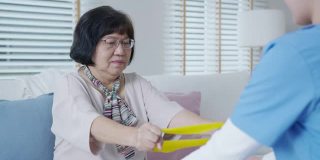 年轻的老年亚洲公民坐在沙发沙发上与家庭护理，辅助生活，搓洗护士医生使用阻力带练习对老年患者在家中进行理疗治疗。