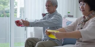 年轻的老年亚洲公民夫妇坐在沙发沙发上配合居家护理，辅助生活，搓洗护士使用阻力带练习为老年患者在家中进行理疗治疗。