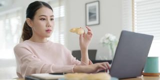 年轻迷人美丽的亚洲女性饿了吃甜甜圈，吃零食吃满嘴看电脑笔记本在家里在忙碌的工作，从家里多任务不健康的饮食生活理念。