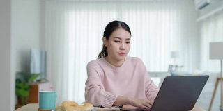 年轻迷人美丽的亚洲女性饿了吃甜甜圈，吃零食吃满嘴看电脑笔记本在家里在忙碌的工作，从家里多任务不健康的饮食生活理念。