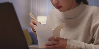 年轻迷人美丽的亚洲女性饿吃方便面中国食品盒带走满嘴看电脑笔记本在家里在忙工作从家里深夜多任务不健康的饮食。