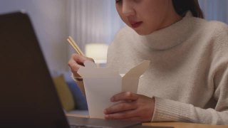 年轻迷人美丽的亚洲女性饿吃方便面中国食品盒带走满嘴看电脑笔记本在家里在忙工作从家里深夜多任务不健康的饮食。视频素材模板下载
