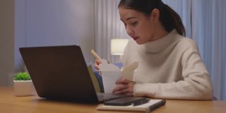 年轻迷人美丽的亚洲女性饿吃方便面中国食品盒带走满嘴看电脑笔记本在家里在忙工作从家里深夜多任务不健康的饮食。