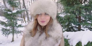 美丽的女人戴着皮帽看着相机在冬天的城市公园里白天在下雪的天气下飘落雪花，慢镜头