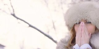 美丽的女人戴着皮帽看着镜头，试图温暖她的手在冬天公园在城市在下雪的天气下，慢镜头