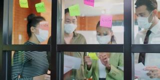 小组头脑风暴的亚洲人把便利贴贴在窗户上。团队合作的商务人士戴上口罩，可以防止新冠病毒感染，在办公室讨论和分享解决问题的想法。