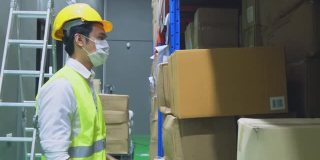 新冠肺炎流行期间，亚洲男性工人戴口罩工作。年轻人带着安全帽、背心搬运产品箱放到物流和出口工厂的货架上。