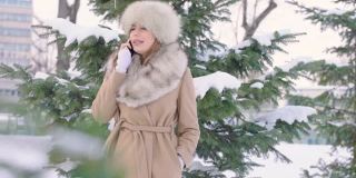 美丽的女人戴着皮帽看相机和使用智能手机在冬天的公园在城市在下雪的天气与飘落的雪，慢镜头