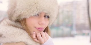 可爱的女人戴着皮帽坐在长椅上，看着镜头在冬天的城市公园里，在下雪的天气里，慢镜头