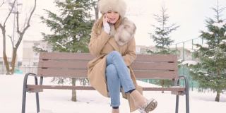 美丽的女人戴着皮草帽坐在长椅上，在冬天的城市公园里使用智能手机在下雪的天气里，慢动作