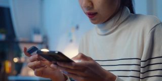 一名年轻的亚洲女性晚上坐在家里的沙发上，一边使用手机一边用信用卡进行网上交易。