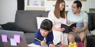 亚洲快乐的女儿正在使用笔记本电脑通过互联网学习，而父母坐在家里的沙发上。电子学习的概念