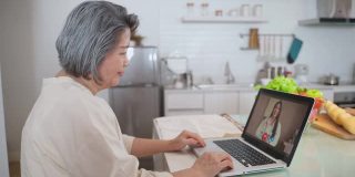 亚洲资深女性视频电话与医生在客厅在家。老年病人咨询全科医生应用电脑。医生和顾问在线和远程医疗的概念。