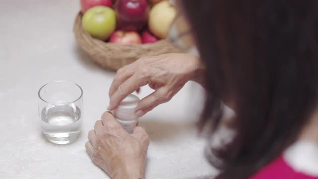 特写亚洲资深妇女服用药物或药丸，并喝了水后，坐在厨房在家里