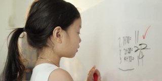快乐的亚洲女孩在家庭教育期间在白板上写下数学科目的问题和答案，呆在家里的概念。