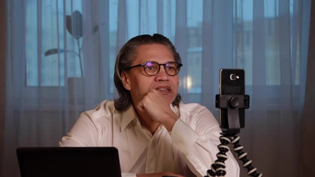 一位50岁的博主戴着眼镜，用智能手机在家录制视频。
