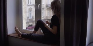 一个女人坐在窗台上，在家里使用智能手机的罕见景象
