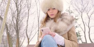 戴着皮帽的漂亮女人坐在公园长椅上，在冬天的城市公园里使用智能手机，在下雪的天气里，特写，慢镜头