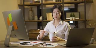亚洲自由职业者精明的商业女性一边吃着方便面，一边在家里的客厅里用笔记本电脑工作。