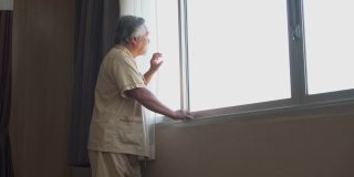 亚洲老年病人站在病房的窗户外。在疗养院里的孤独老人