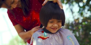 亚洲母亲正在修剪她的女儿的头发在早上在花园里。在COVID-19或冠状病毒大流行期间的国内