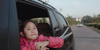 在去学校的路上，小女孩从车窗里伸出手，笑着，微笑着。亚洲小女孩微笑着，示意把车伸出来。孩子们在车里可以看到街景，放松心情。家庭在汽车的概念。