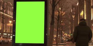 人行道上的绿色屏幕广告牌，靠近繁忙的街道，供人们行走。汽车在晚上行驶。
