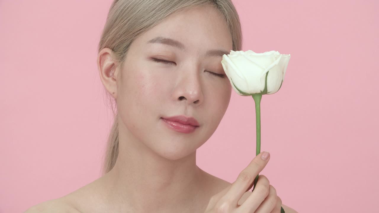 一个年轻，美丽的亚洲女人的特写肖像与金发握着，闻白色的花，然后看着相机在粉红色的背景。博主与美丽，健康或销售网上概念。